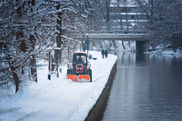 Wall Mural - Tractor with snow plow and rotating brush sweeping snow from foopath on embankment in park. Red tractor with scoop and automated brush removes snow. Municipal road sweeping vehicle with plow