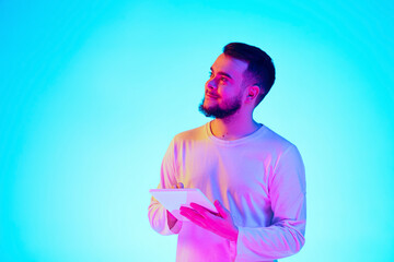 Scrolling tablet. Caucasian man's portrait with device on blue studio background in neon light. Beautiful male model. Concept of human emotions, facial expression, sales, ad. Copyspace for ad. Close