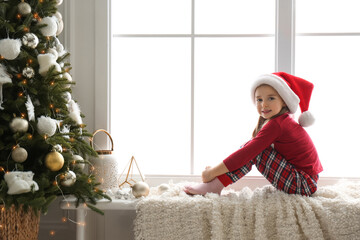 Wall Mural - Cute little girl in Santa hat on window sill near Christmas tree at home