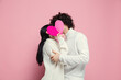 © master1305 - Young, beautiful couple in love on pink studio background. Saint Valentine's Day, love, relationship and human emotions concept. Copyspace. Young man and woman hiding behind the heart shaped card.