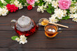 Steamed glass teapot, elegant cup with tea and petals, honey dipper on a wooden table among flowering jasmine branches and roses. Outdoor, picnic, brunch. Top view