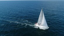 Aerial View. A White Sailboat Is Moving Fast On The Waves Of The Sea. Flying Above A Boat In Full Sailing Gear. Close Hauled Sailing In Ocean. Yacht In Windy And Sunny Day. Drone Shot.
