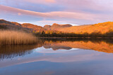 Fototapeta Morze - Mirrored reflections in calm lake with golden light from sunrise on mountain tops. Elterwater, Lake District, UK.