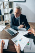 © LIGHTFIELD STUDIOS - Mature investor looking at businesspeople while holding documents with charts near laptop in office