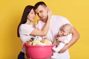 Tired troublesome parents crying newborn baby and basin full of clean clothing, have much domestic work, posing isolated on yellow background, fall in love sleepy parents hug and having minute rest.