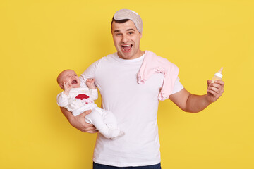 Father feeding baby with bottle isolated over yellow background, excited handsome dad with infant on hands, guy yelling with happy expression.