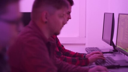 Male programmers sitting in the office at computers start the working day and type quickly on the keyboard. Pink light in a dark room. Fashionable lighting.