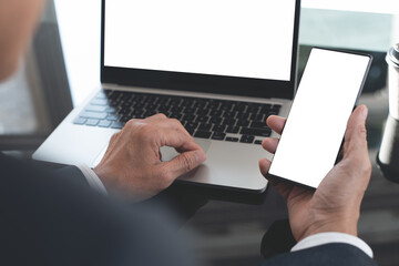 Canvas Print - Mockup image, business man hand holding black black mobile smart phone working on laptop computer at office