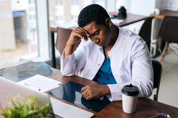 Poster - Tired African American black man doctor in white coat working on laptop, thinking about problem, touching forehead. Concept of medicine and health care.