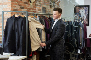 A young man in a suit is choosing a shirt or hoodie in a clothing store. A male customer in a boutique.