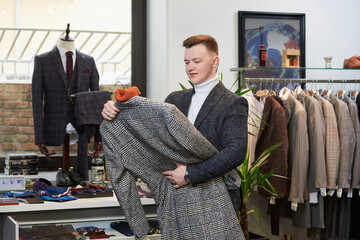 A young man in a white turtleneck sweater and a suit is choosing a coat on a hanger in a clothing store. A male customer in a boutique.