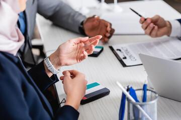 Sticker - cropped view of arabian businesswoman pointing with hand near multicultural business partners and digital translators, blurred background