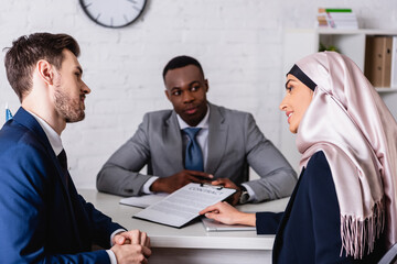 Sticker - smiling arabian businesswoman pointing with hand at contract during discussion with multiethnic business partners, blurred background