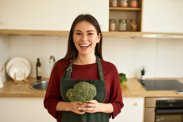 Excited positive young housewife holding organic broccoli, standing in kitchen, laughing, cooking vegetarian meal for lunch. Pretty girl in apron making healthy vegan raw dinner using fresh vegetables