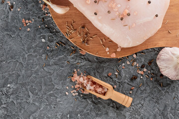 Raw chicken fillet on a cutting board on a gray stone texture, next to sea salt, pepper peas and garlic. Top view, copy space, flat lay