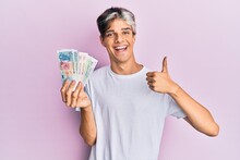 Young Hispanic Man Holding Singapore Dollars Banknotes Smiling Happy And Positive, Thumb Up Doing Excellent And Approval Sign