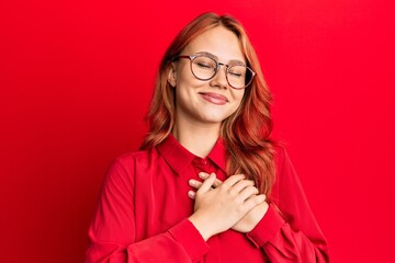 Wall Mural - Young beautiful redhead woman wearing casual clothes and glasses over red background smiling with hands on chest with closed eyes and grateful gesture on face. health concept.