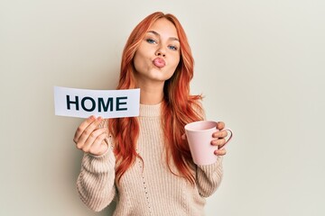 Wall Mural - Young redhead woman drinking a cup of coffee holding home text looking at the camera blowing a kiss being lovely and sexy. love expression.