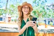 © Krakenimages.com - Young beautiful blonde woman on vacation wearing summer hat and glasses smiling happy. Standing with smile on face using smartphone at street of city.