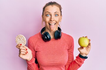 Poster - Beautiful caucasian sports woman holding green apple and rice crackers sticking tongue out happy with funny expression.