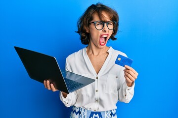 Wall Mural - young brunette woman wearing business style holding laptop and credit card angry and mad screaming f