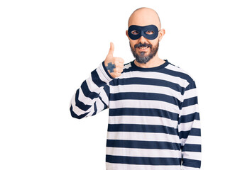 Poster - Young handsome man wearing burglar mask doing happy thumbs up gesture with hand. approving expression looking at the camera showing success.