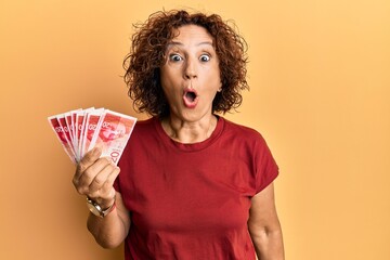 Poster - Beautiful middle age mature woman holding 20 israel shekels banknotes scared and amazed with open mouth for surprise, disbelief face