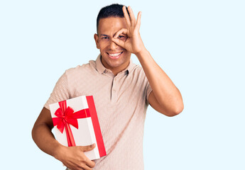 Wall Mural - Young handsome latin man holding gift smiling happy doing ok sign with hand on eye looking through fingers