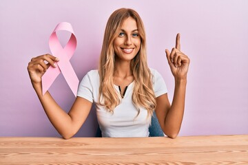 Sticker - Beautiful blonde young woman holding pink cancer ribbon surprised with an idea or question pointing finger with happy face, number one