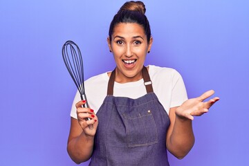 Poster - Young beautiful brunette woman wearing apron holding whisk celebrating achievement with happy smile and winner expression with raised hand