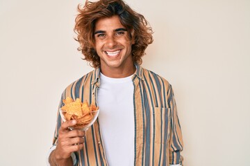 Young hispanic man holding nachos potato chips looking positive and happy standing and smiling with a confident smile showing teeth