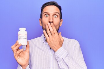 Canvas Print - Young handsome man holding jar of medicine pills standing over isolated purple background covering mouth with hand, shocked and afraid for mistake. Surprised expression