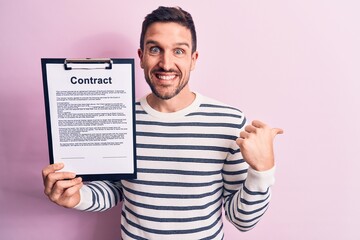 Wall Mural - Young handsome man holding clipboard with contract document paper over pink background pointing thumb up to the side smiling happy with open mouth