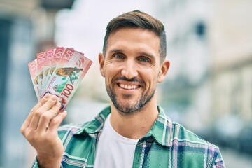 Poster - Young caucasian man smiling happy standing holding new zealand dollars banknotes at the city.