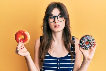 Sticker - Young beautiful caucasian girl holding tasty colorful doughnuts clueless and confused expression. doubt concept.