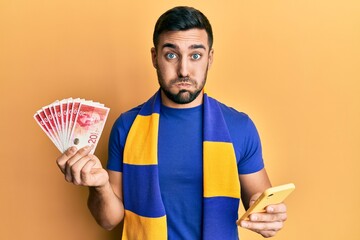 Canvas Print - Young hispanic man football supporter using smartphone holding israel shekels banknotes puffing cheeks with funny face. mouth inflated with air, catching air.