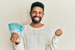 Handsome hispanic man with beard holding 100 brazilian real banknotes screaming proud, celebrating victory and success very excited with raised arm