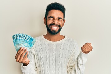 Wall Mural - Handsome hispanic man with beard holding 100 brazilian real banknotes screaming proud, celebrating victory and success very excited with raised arm