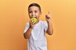 © Krakenimages.com - Adorable latin toddler smiling happy eating green apple looking to the camera over isolated yellow background.