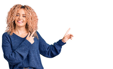 Young blonde woman with curly hair wearing casual winter sweater smiling and looking at the camera pointing with two hands and fingers to the side.