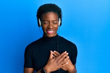 Poster - Young african american girl listening to music using headphones smiling with hands on chest with closed eyes and grateful gesture on face. health concept.