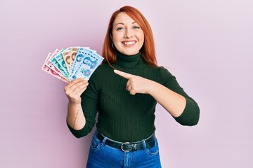 Sticker - Beautiful redhead woman holding yuan chinese banknotes smiling happy pointing with hand and finger