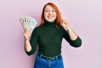 Sticker - Beautiful redhead woman holding united kingdom pounds smiling with an idea or question pointing finger with happy face, number one