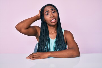 Sticker - African american woman with braids wearing casual clothes sitting on the table confuse and wonder about question. uncertain with doubt, thinking with hand on head. pensive concept.