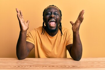 Canvas Print - Handsome young black man wearing casual clothes sitting on the table celebrating mad and crazy for success with arms raised and closed eyes screaming excited. winner concept