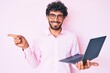 © Krakenimages.com - Handsome young man with curly hair and bear working using computer laptop smiling happy pointing with hand and finger to the side