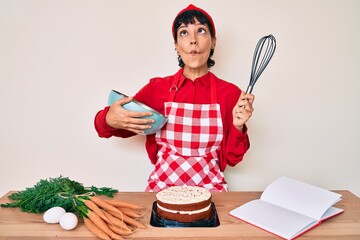 Beautiful brunettte woman cooking carrot cake using whisk making fish face with mouth and squinting eyes, crazy and comical.