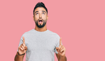 Canvas Print - Young man with beard wearing casual grey tshirt amazed and surprised looking up and pointing with fingers and raised arms.