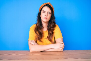 Young beautiful brunette woman sitting on the table over blue background skeptic and nervous, disapproving expression on face with crossed arms. negative person.