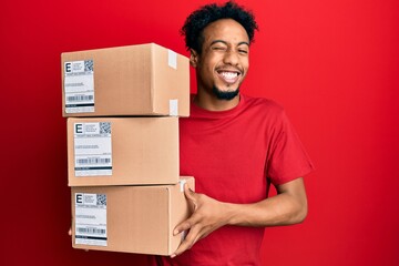 Young african american man with beard holding delivery packages winking looking at the camera with sexy expression, cheerful and happy face.